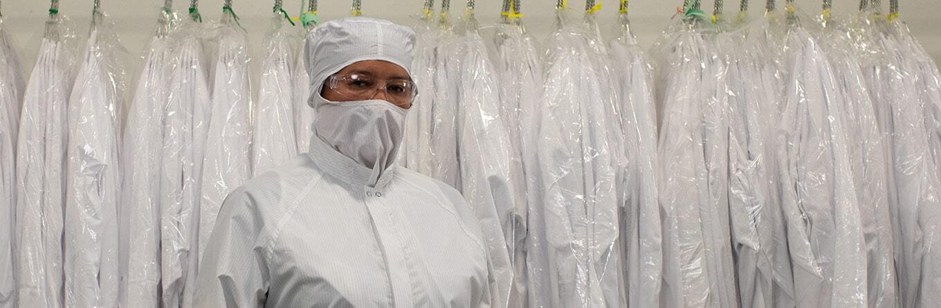 A cleanroom technician stands in front of a rack of white protective garments while wearing a hygienically cleaned and processed garment and clear safety glasses provided by UniFirst as part of a custom uniform and facility service program.