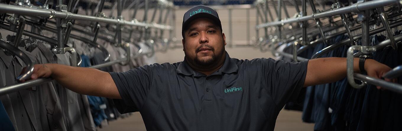 A UniFirst production team partner wearing a Micro Piqué Polo Shirt with UniFirst logo and a baseball hat with UniFirst logo stands with sorted and hygienically laundered workwear in a UniFirst ISO-9001 certified processing facility.