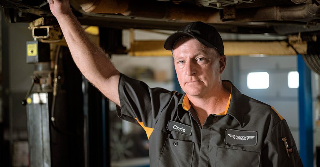 Uniformed automotive mechanic stands under vehicle in lift.