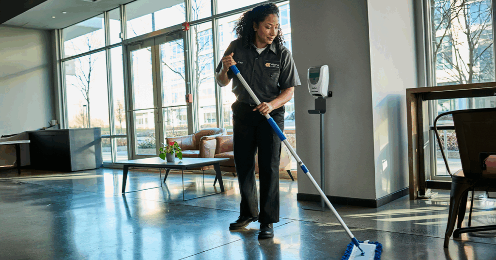 Woman in office wearing UniFirst workwear attire, mopping the floor with a dry mop.