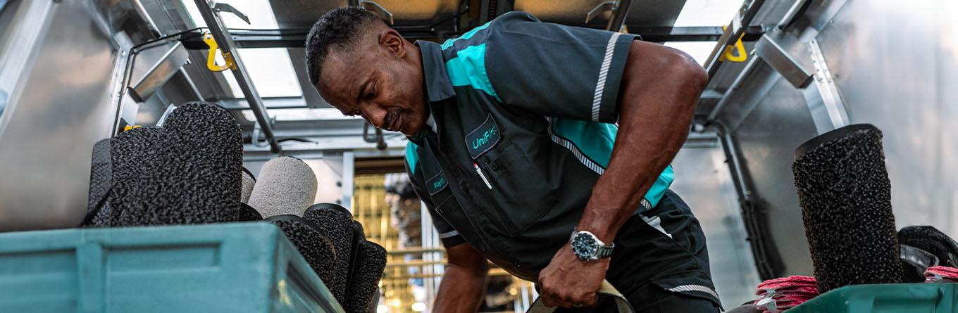 A UniFirst Route Service Representative wearing a Spotlite MV Short Sleeve Enhanced Visibility Polo Shirt carries a freshly laundered Great Impressions<sup>®</sup> 2.0 Walk-Off Mat, exclusively made by UniFirst in the USA at one of our ISO-9001 certified manufacturing plants, to a UniFirst facility services program customer.
