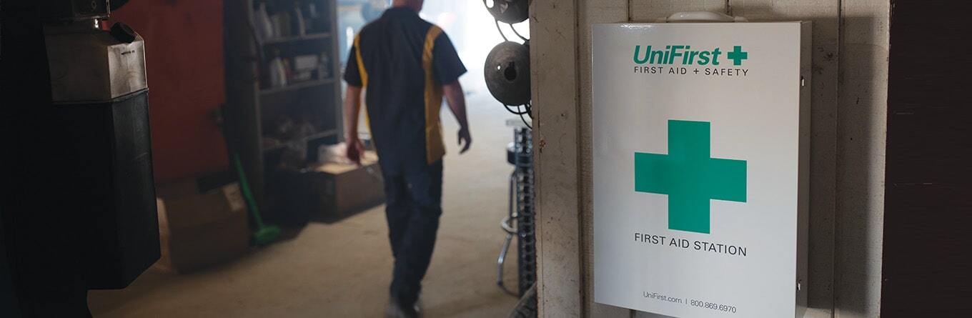 A UniFirst First Aid & Safety First Aid Station cabinet is placed on the wall of a automotive repair shop.